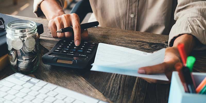 Someone calculates figures on a calculator while reviewing paperwork on a wooden desk.