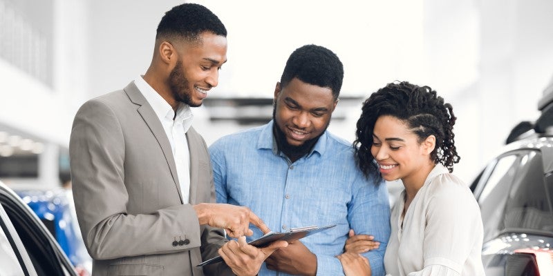 Car salesman pointing to a document on a clipboard for a smiling couple in a car showroom.