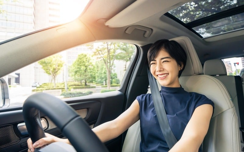 Smiling Asian woman driving a modern car with a panoramic sunroof, sunlight streaming in from the top left.