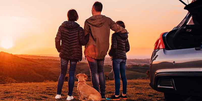 Family and dog stand by a car with an open trunk, watching the sunset over hills.