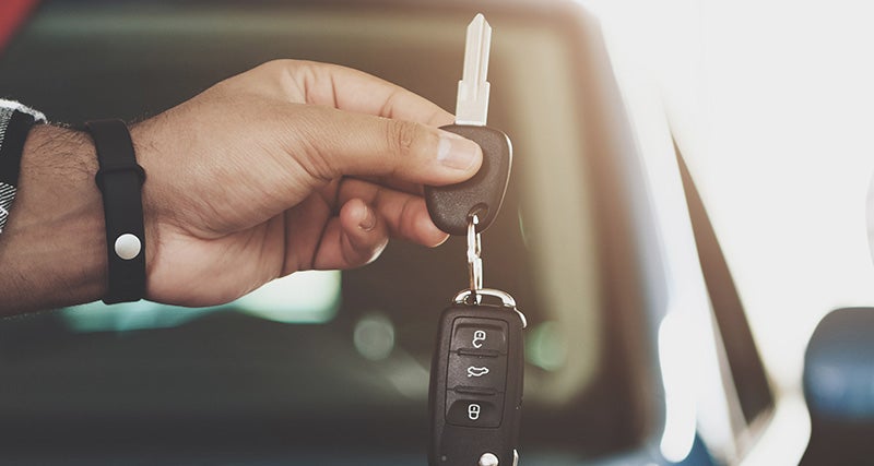 A hand, wearing a dark wristband, holds up a set of car keys with a remote fob against a blurred background.