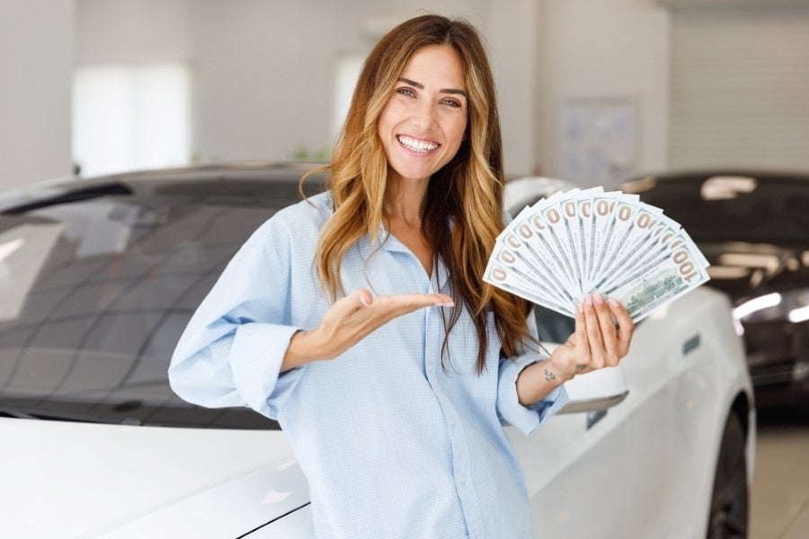 A smiling woman holds a fan of one hundred dollar bills in front of a black car.