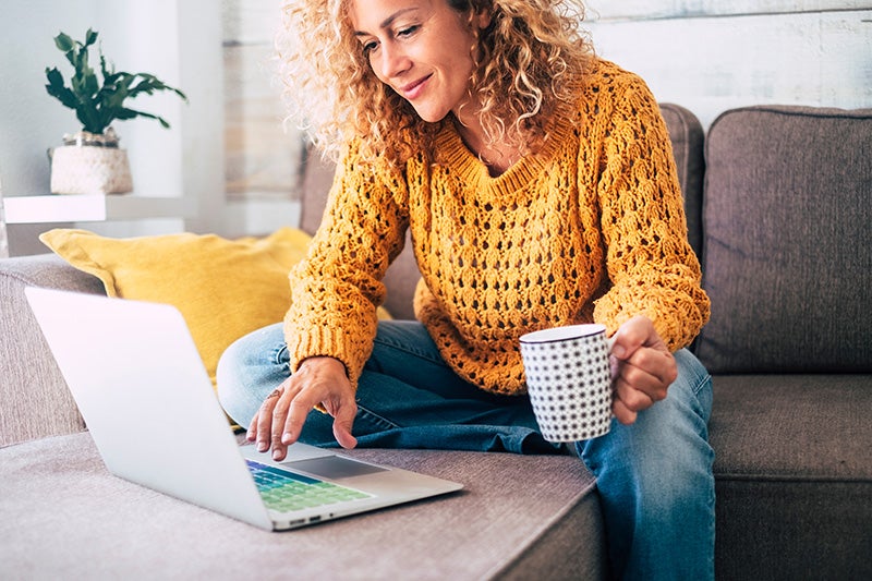 A woman with curly hair smiles while using a laptop and holding a mug on a sofa.